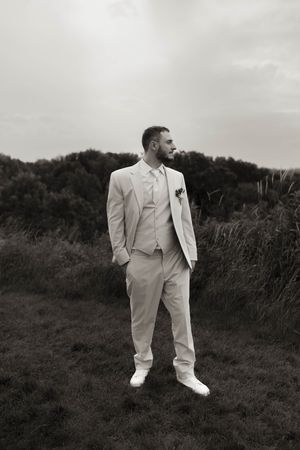 Groom in white suit poses dramatically against natural landscape.