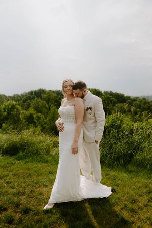 Newlyweds share intimate moment on green hillside in matching white attire.