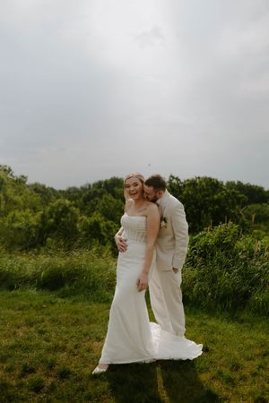 Couple in white wedding attire embraces on lush green hillside.