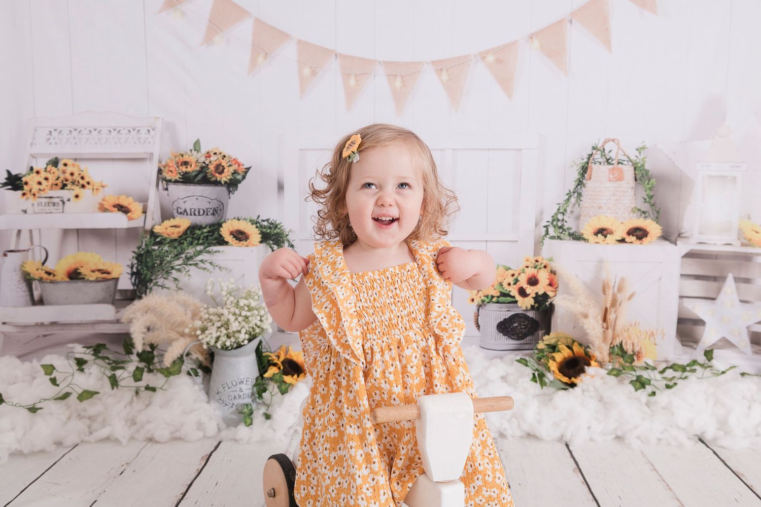 A young child in a yellow dress smiles while sitting on a rustic floor with floral decorations and pink bunting overhead.