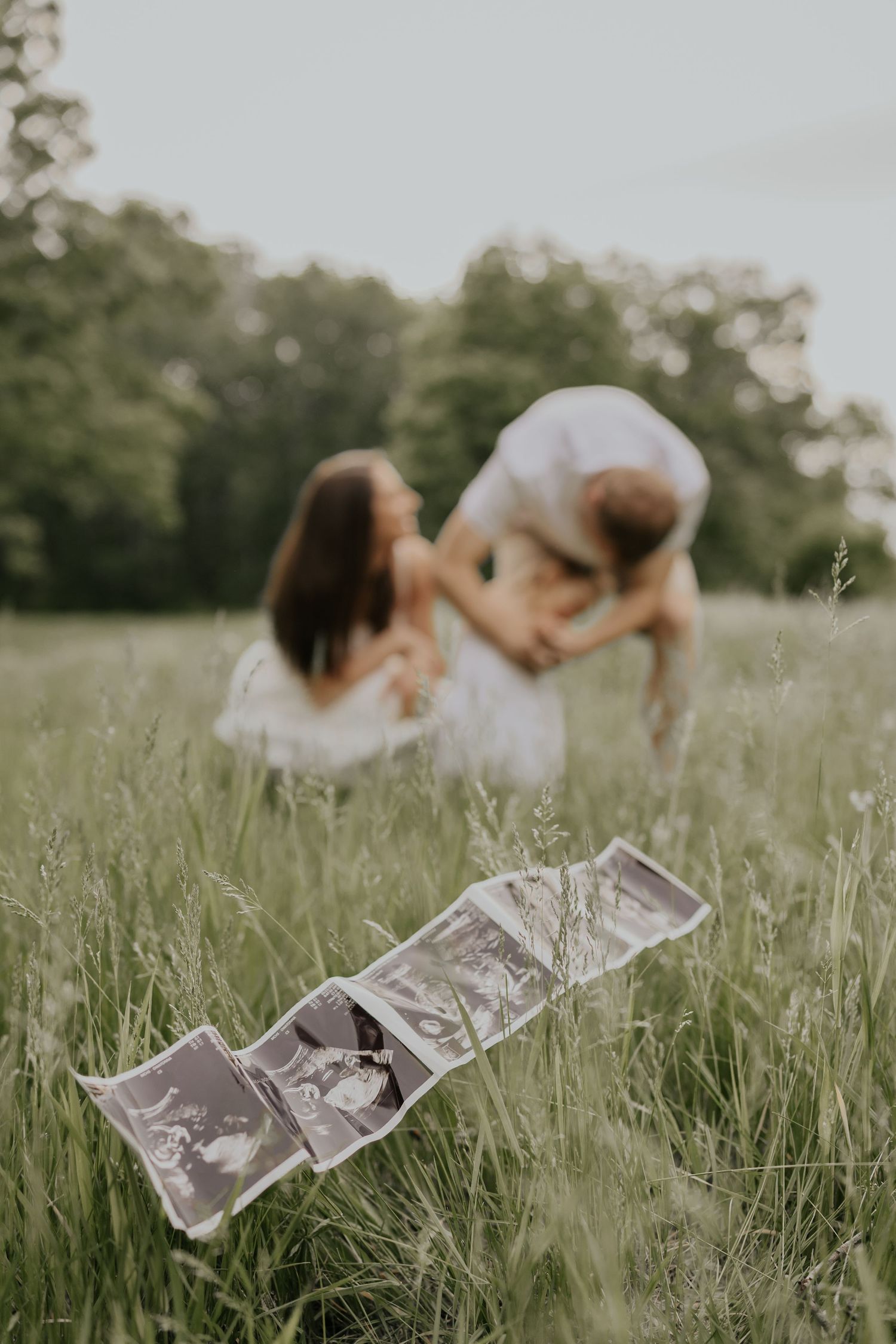 Ultrasound photos displayed in tall grass while a couple embraces in a soft focus background during pregnancy announcement shoot.