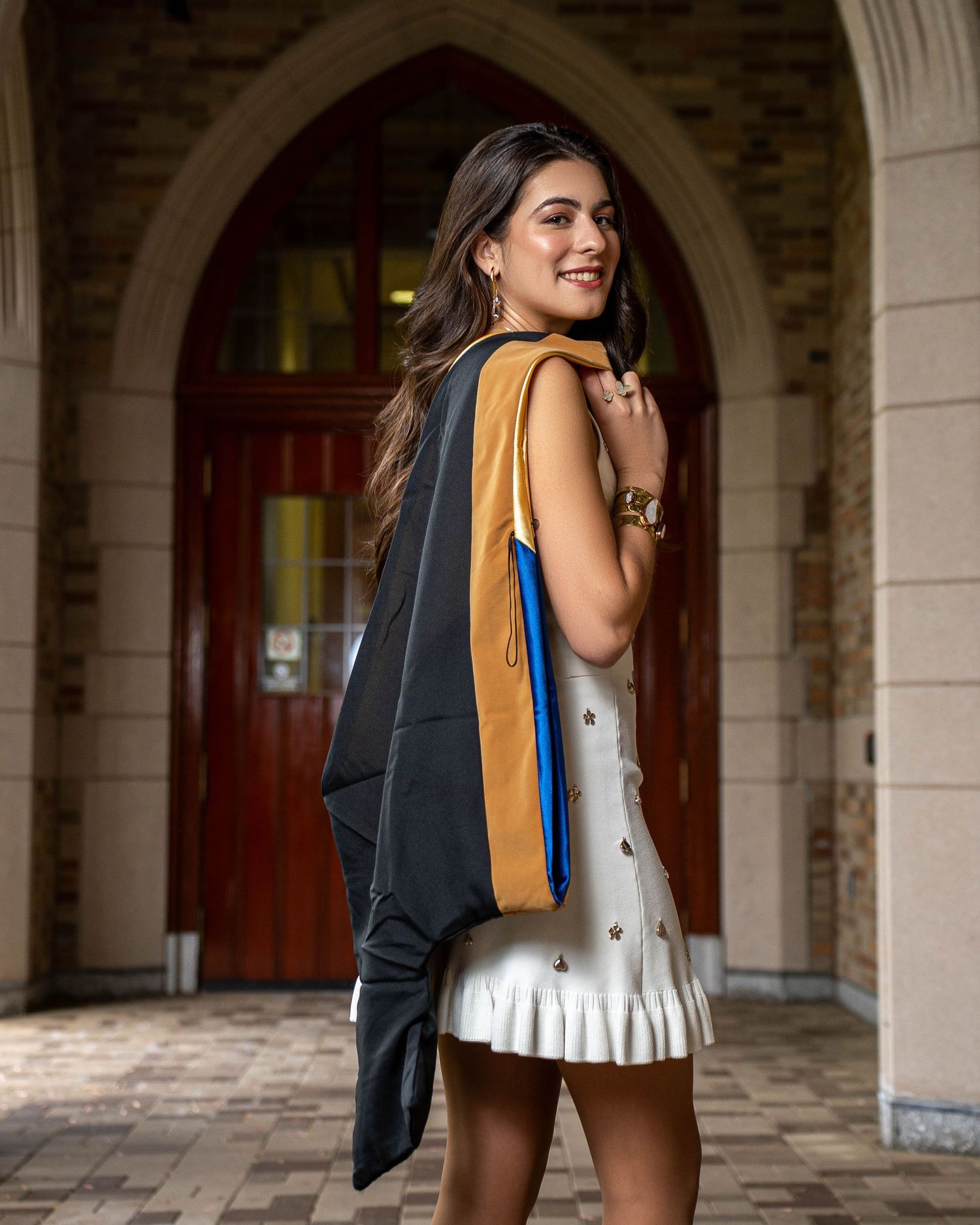 A person wearing a white dress and carrying a blue and orange tote bag stands in an archway with stone columns.