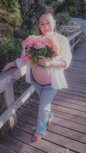 A child in jeans holds a bouquet of pink flowers while standing on a wooden boardwalk in nature.