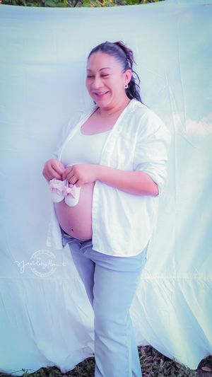A pregnant woman in a white cardigan and jeans stands against a light backdrop holding tiny baby shoes and smiling warmly.