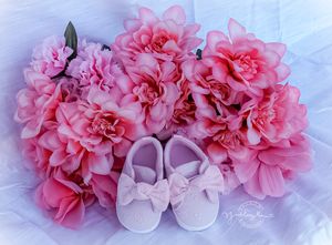 Pink baby shoes with bows surrounded by blooming pink carnation flowers on a light blue background.