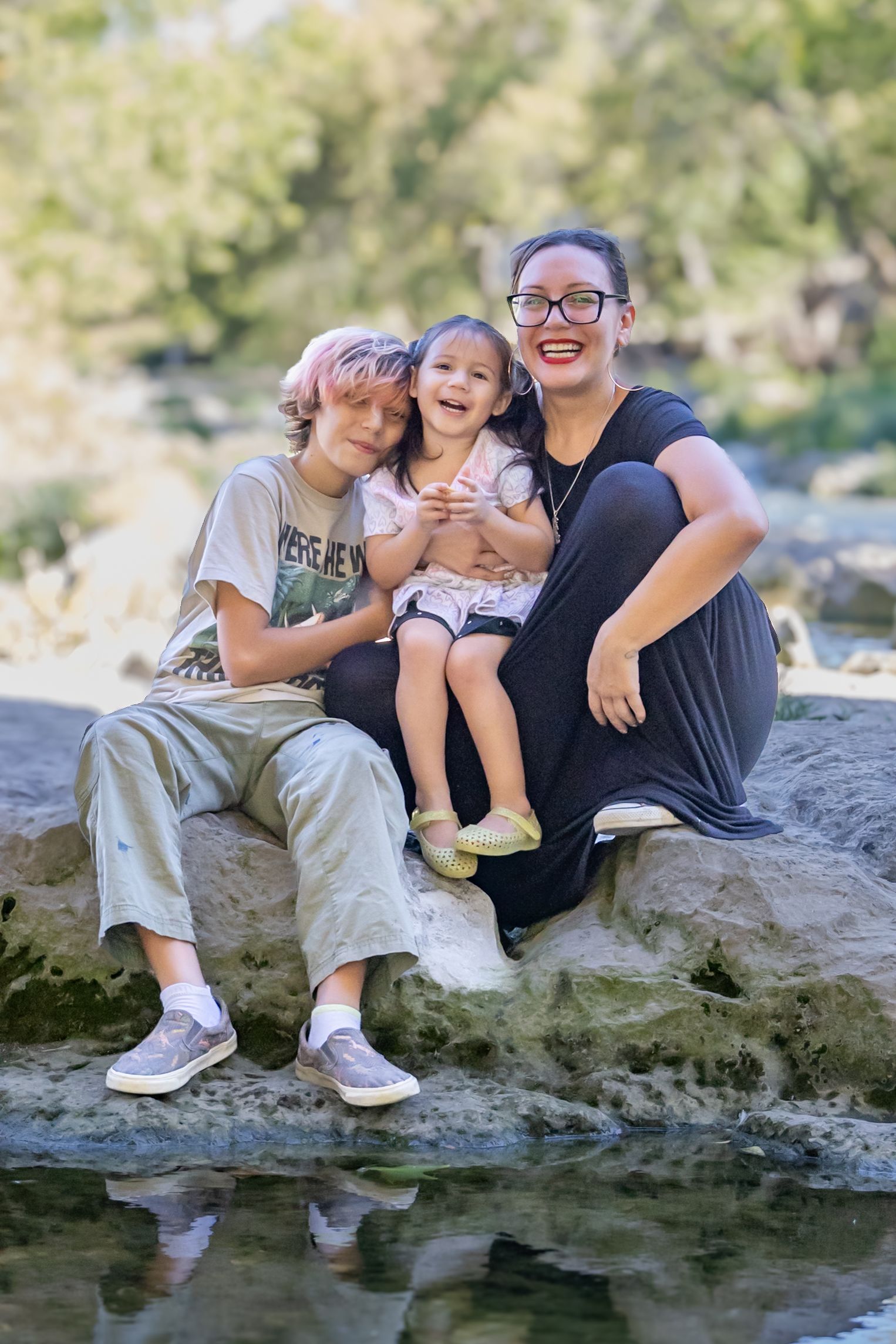 A happy family sits together on a rock by a stream on a sunny day, sharing joyful smiles and laughter in nature.