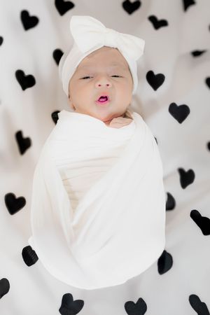 Black heart pattern wall provides backdrop for artful baby photography series in white swaddling.