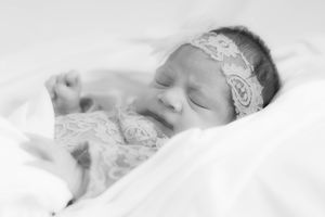 A newborn baby wearing lace headband and dress sleeps peacefully on white bedding in black and white photography.