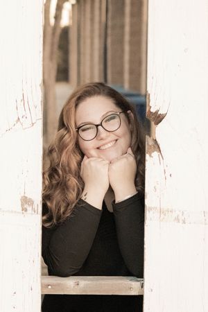Portrait of a person with curly brown hair and glasses posing in a window frame with their hands under their chin.