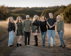 A family group of seven people poses together on a dirt path with trees in the background at sunset.
