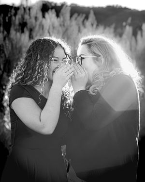 Two people share a secret moment together in black and white, leaning close and laughing against backlit scenery.