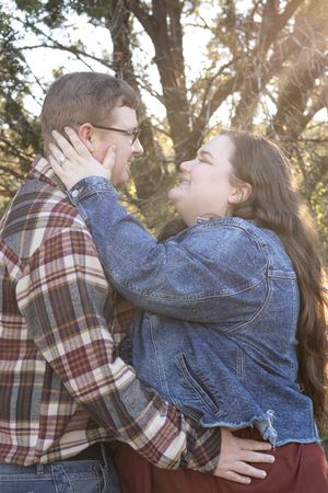 A couple in casual denim and plaid embraces lovingly outdoors with sunlight filtering through trees in the background.