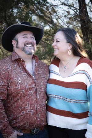 A joyful couple shares a laugh outdoors, the person in a cowboy hat wearing western attire and the other in a striped sweater.