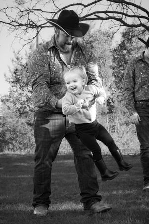 Black and white portrait of a person in a cowboy hat playfully lifting a laughing toddler outdoors under tree branches.