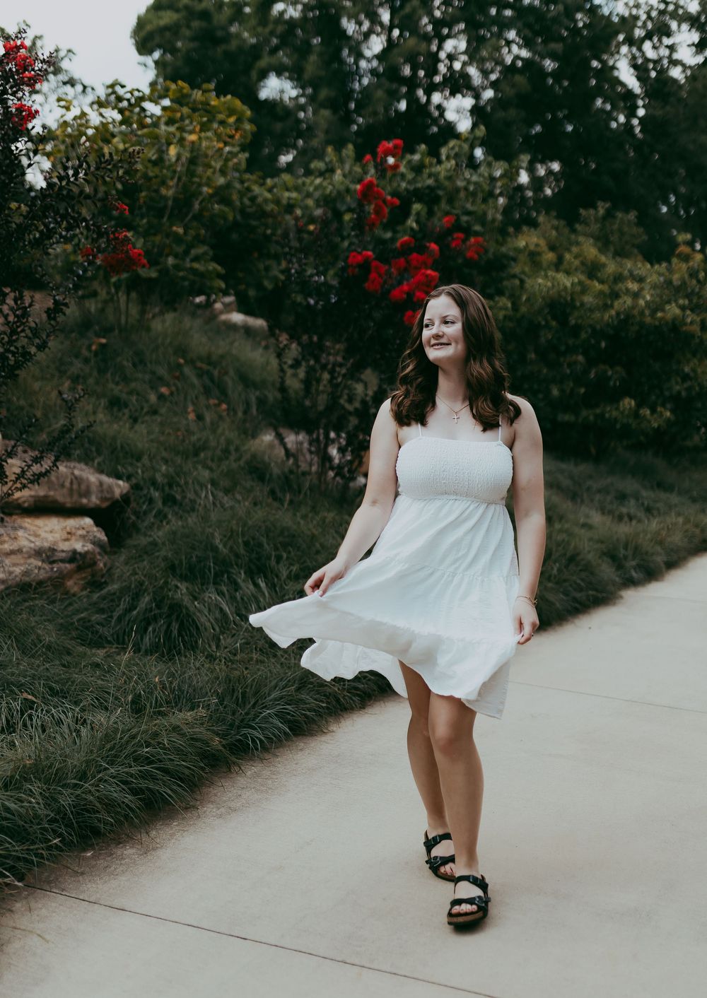 A white flowy sundress flows in the breeze along a garden path with red flowers and greenery in the background.