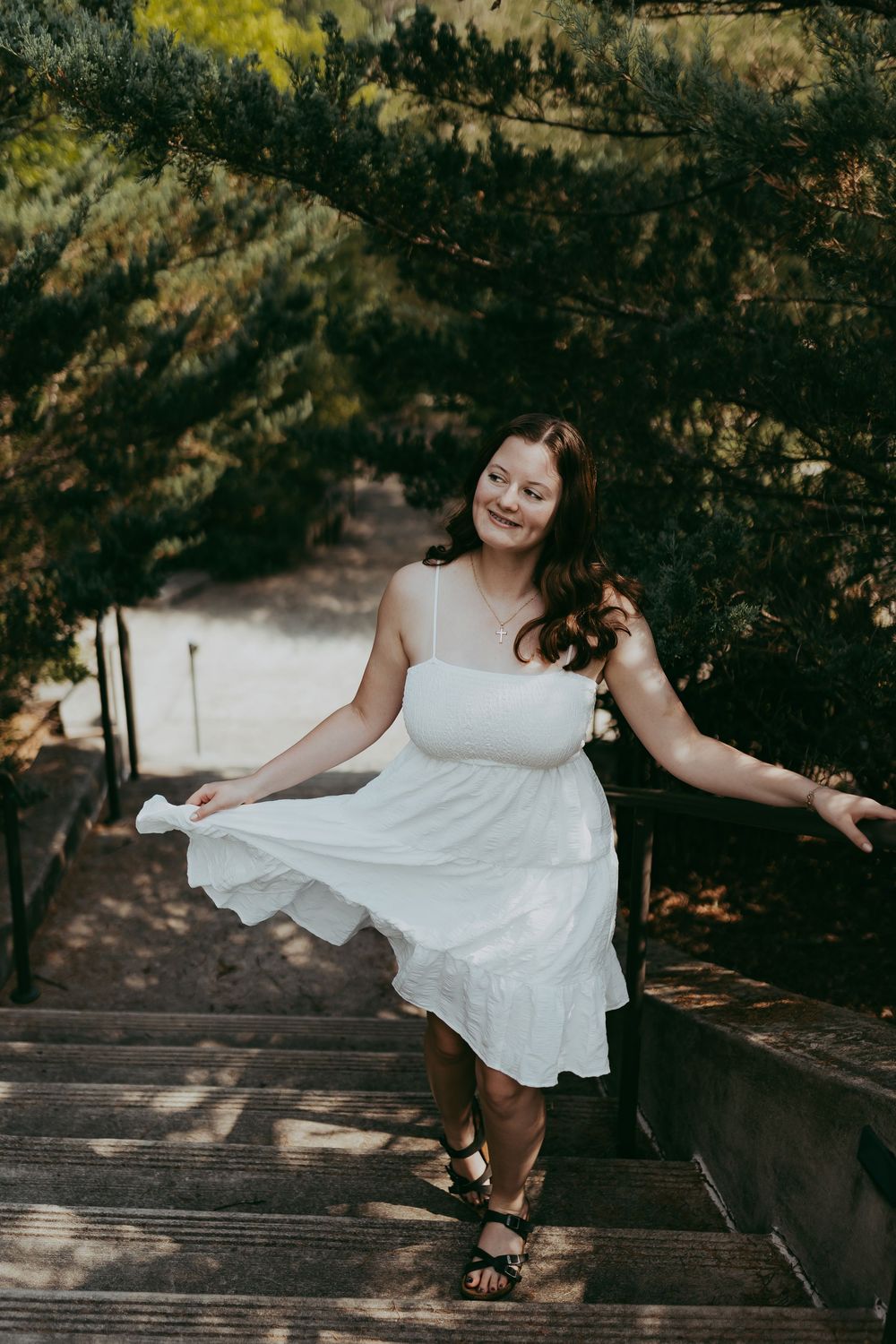 A person in a flowy white dress strikes a playful pose on wooden stairs surrounded by lush greenery.