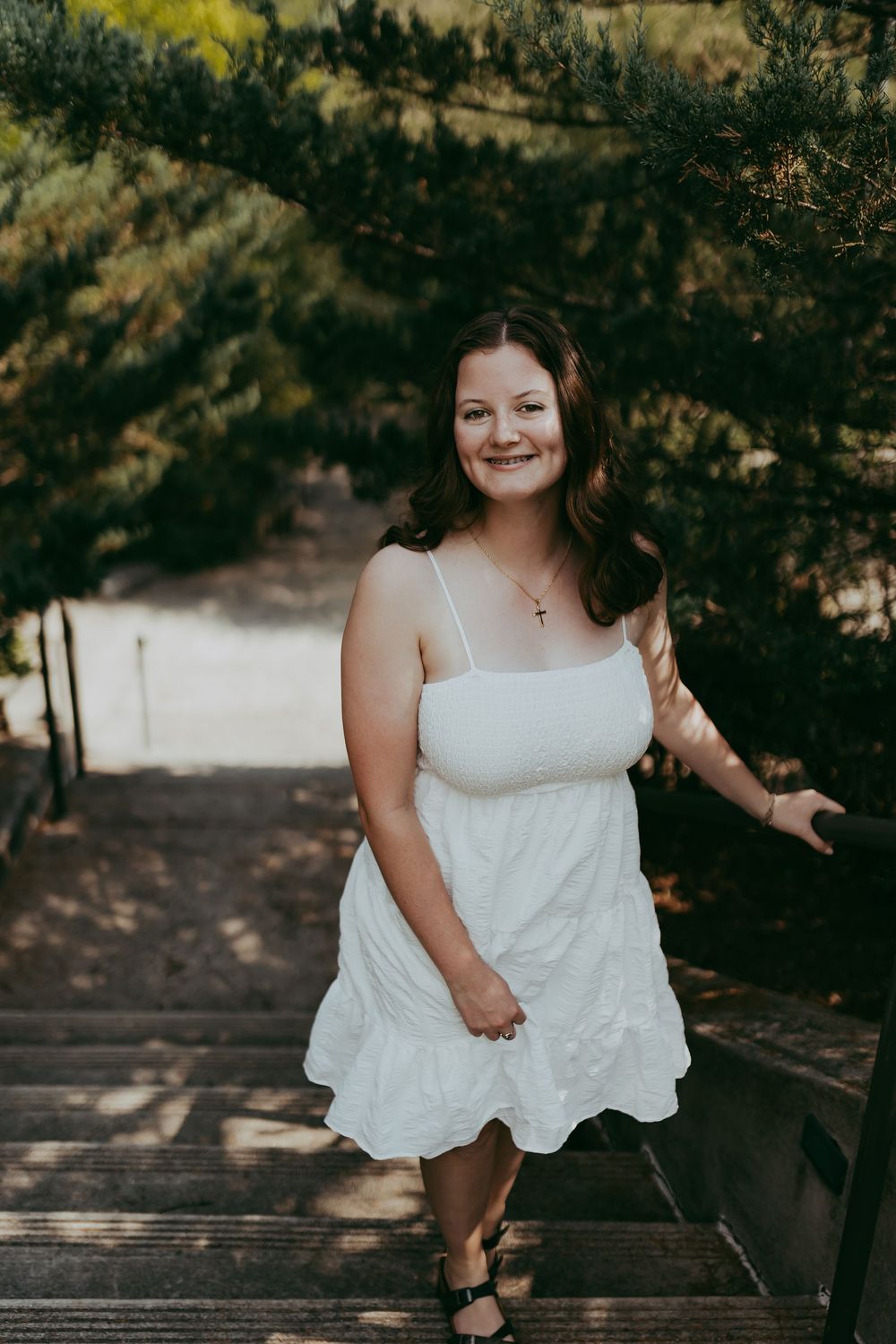 A person in a white sundress stands on wooden stairs surrounded by pine trees on a sunny day.