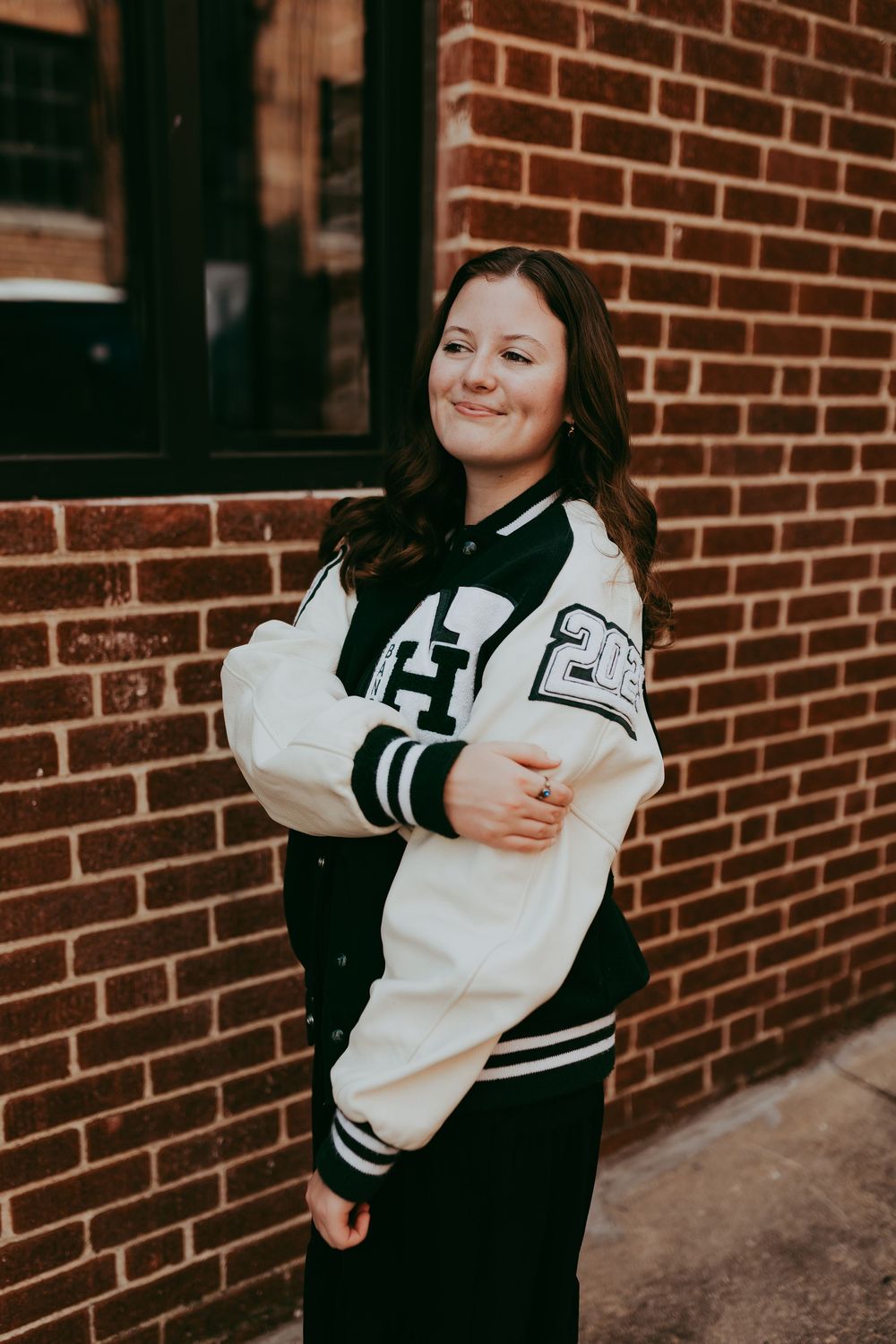 Person wearing a black and white varsity letterman jacket poses against brick wall outdoors.