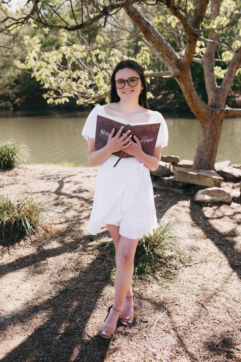 Someone in a white summer dress reads a book while standing by a lake on a sunny day under a tree.