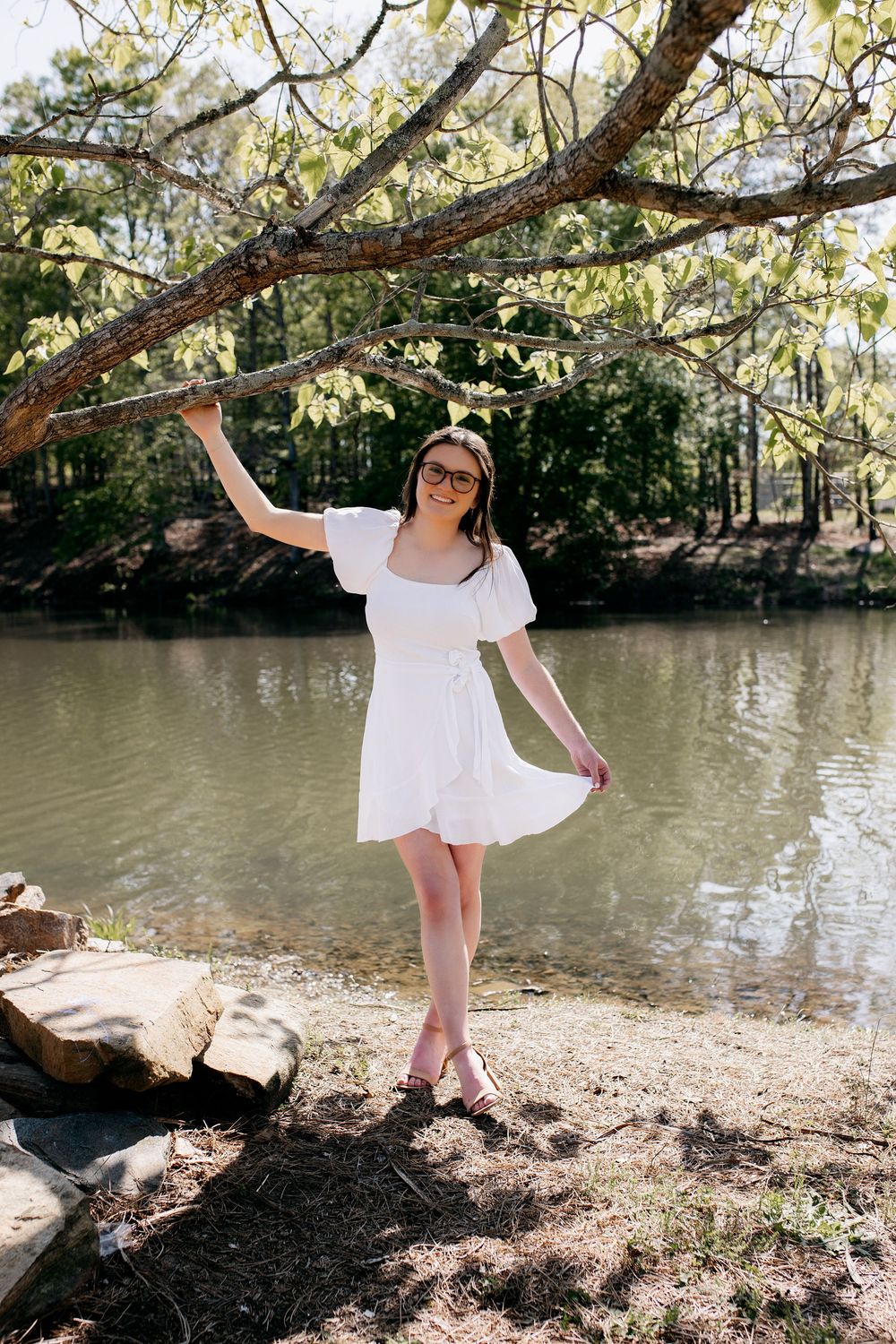 A person in a white summer dress poses gracefully by a peaceful pond beneath overhanging tree branches on a sunny day.