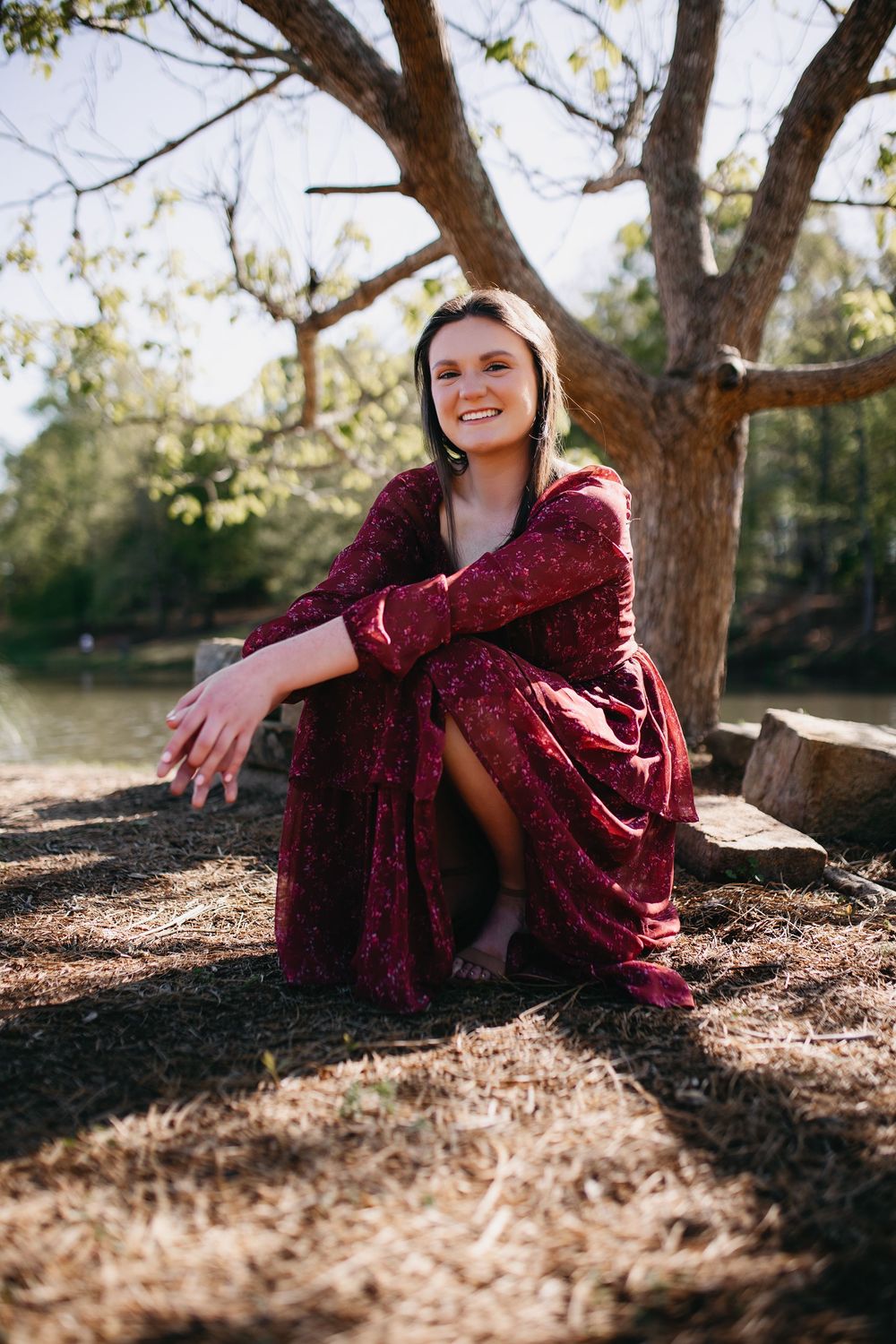 Someone wearing a burgundy sequined dress sits on fallen leaves under a large tree on a sunny autumn day.