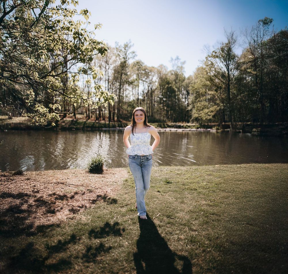 Person stands by a serene pond surrounded by tall trees on a sunny day in a natural outdoor setting.