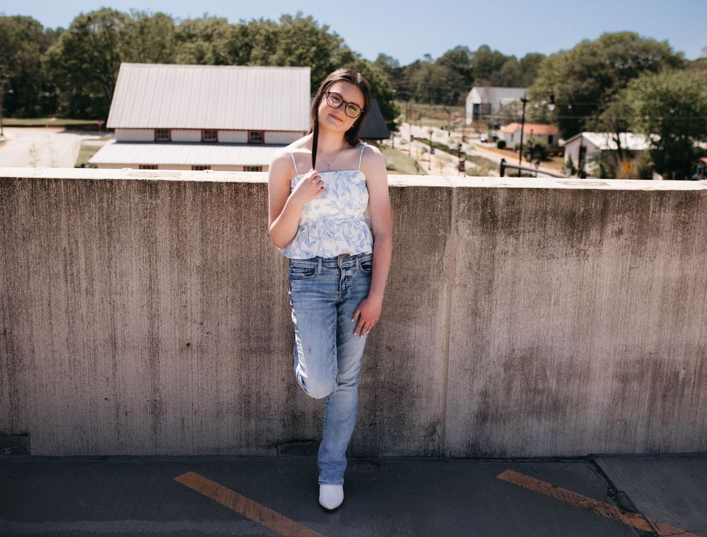 Person in white top and distressed jeans leans against concrete wall on rooftop with buildings and trees in background.