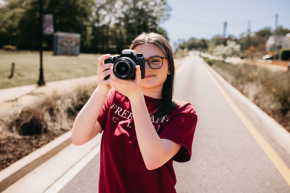 Photographer in burgundy t-shirt takes picture with DSLR camera on empty suburban road on sunny day.
