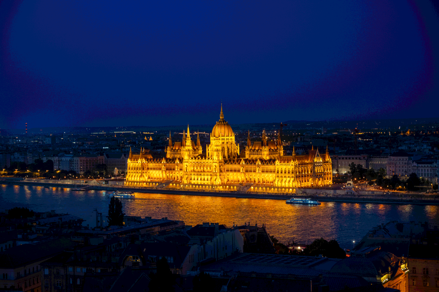 Illuminated Hungarian Parliament Building glows golden against dark blue night sky along Danube River in Budapest.