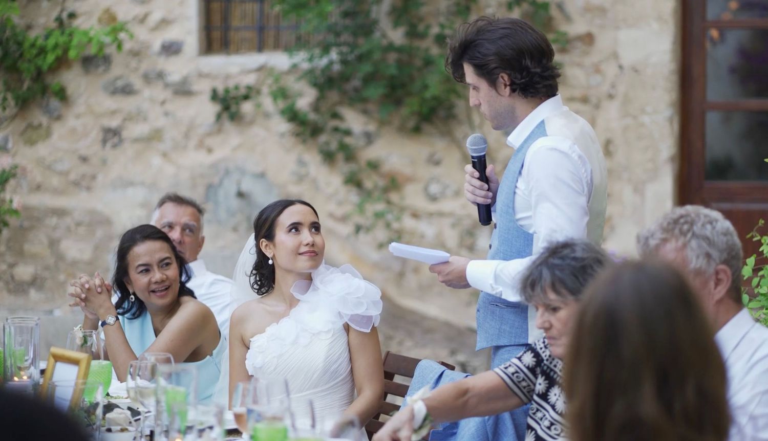 Man giving a speech at an outdoor wedding, while the bride listens with a smile surrounded by guests.