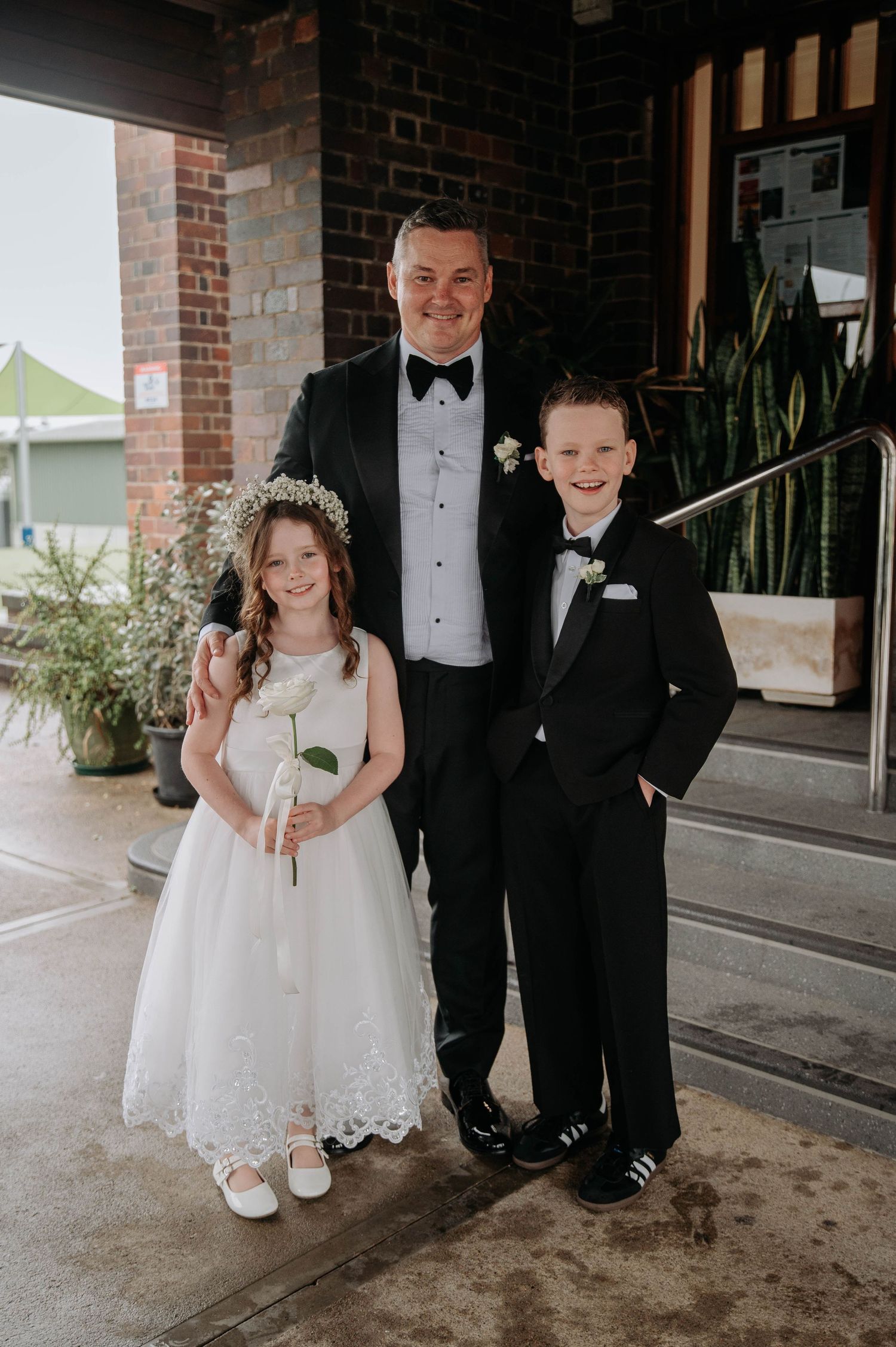 Wedding party photos showing flower girls in white lace dresses with tuxedo-clad companions on a covered brick walkway.