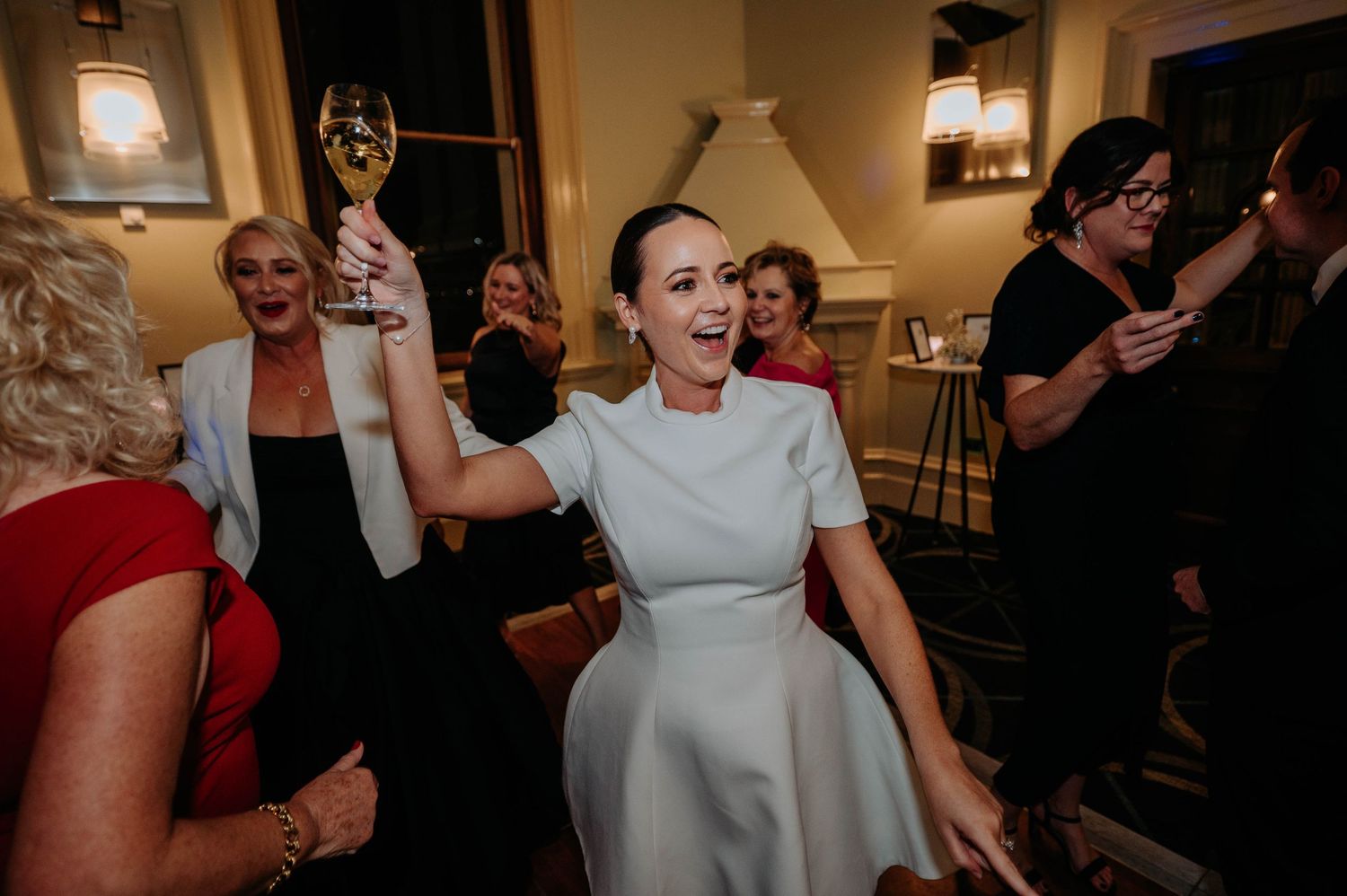 A lively wedding reception scene showing guests dancing and celebrating in a warmly lit indoor venue.