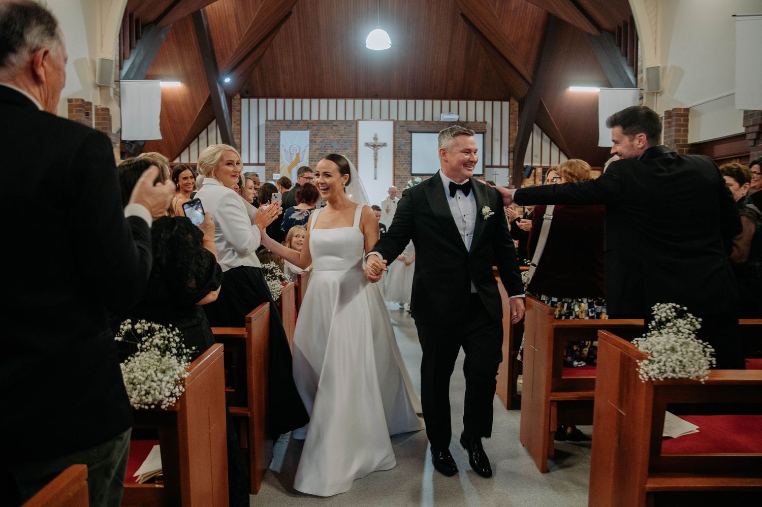 Newlywed couple joyfully exits church ceremony while guests celebrate with flower petals and bubbles in wooden pew-lined aisle.