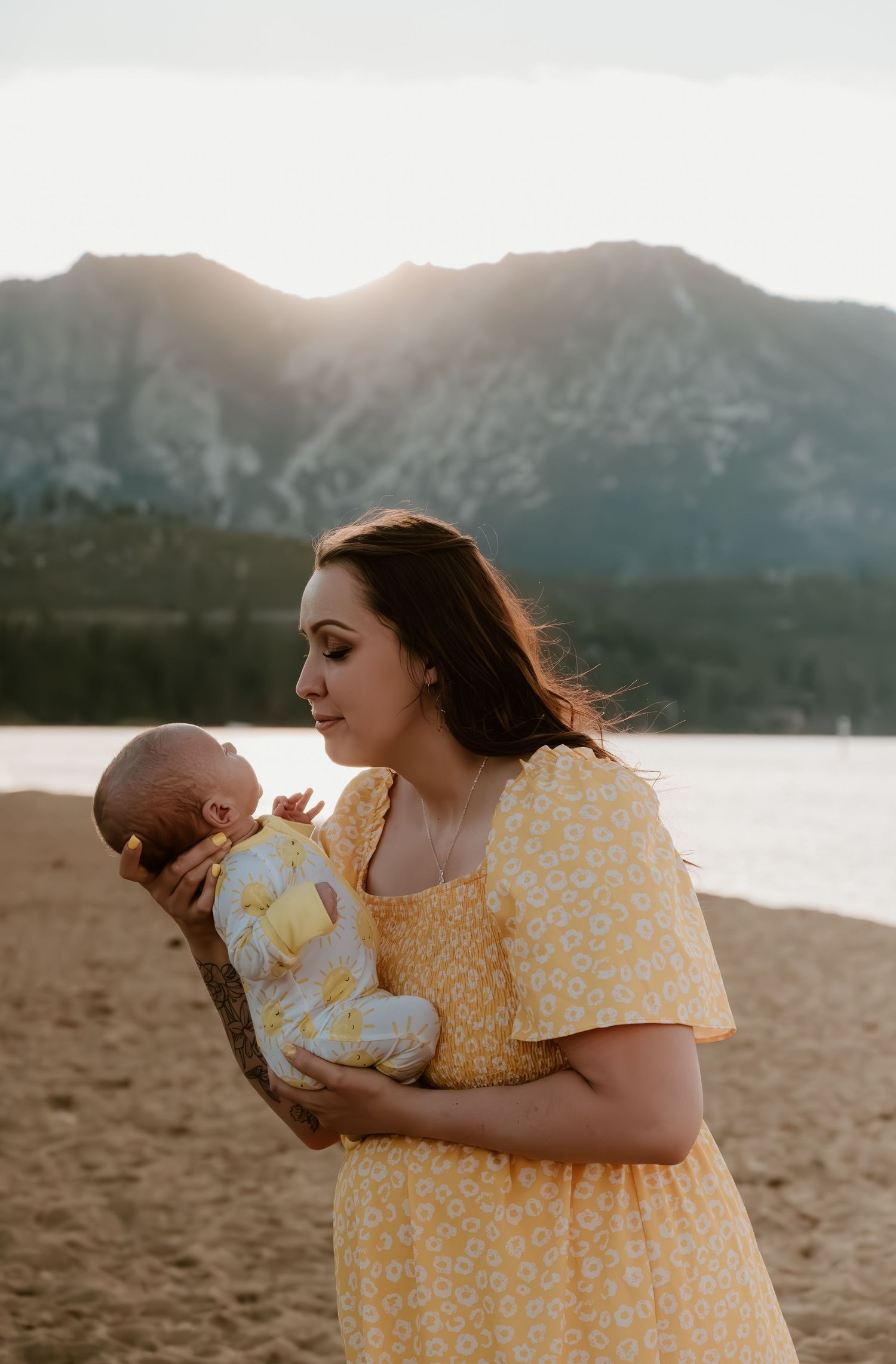 Backlit by sunset over mountain lake, figure in yellow floral dress tenderly holds infant on sandy beach.