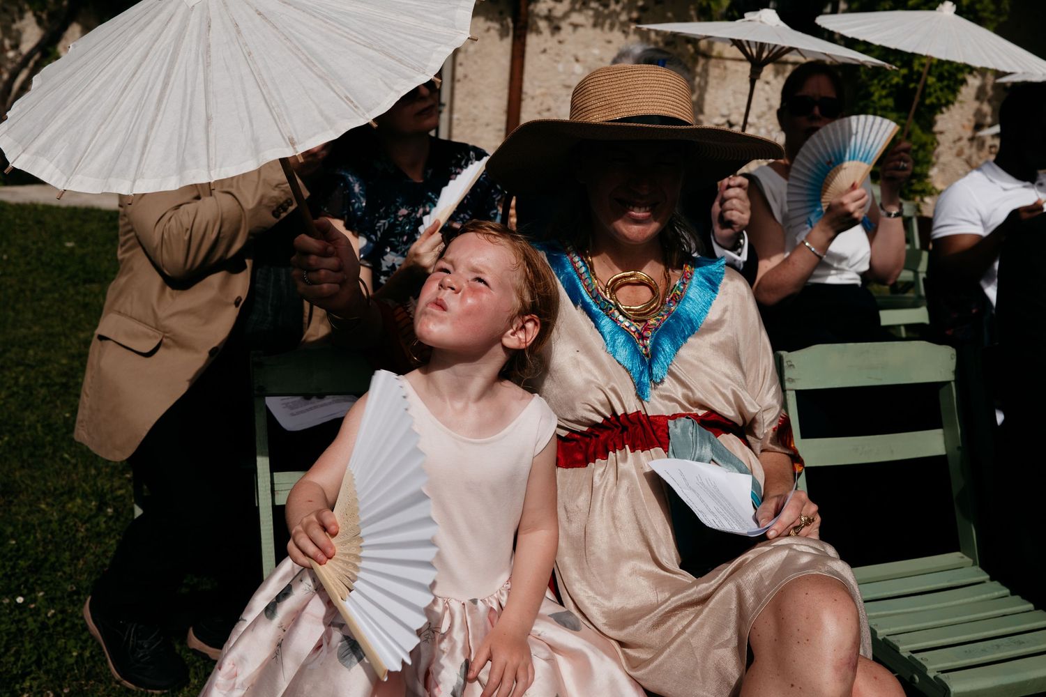 Wedding guests shelter from sun with parasols and fans while seated on green wooden chairs at outdoor ceremony.