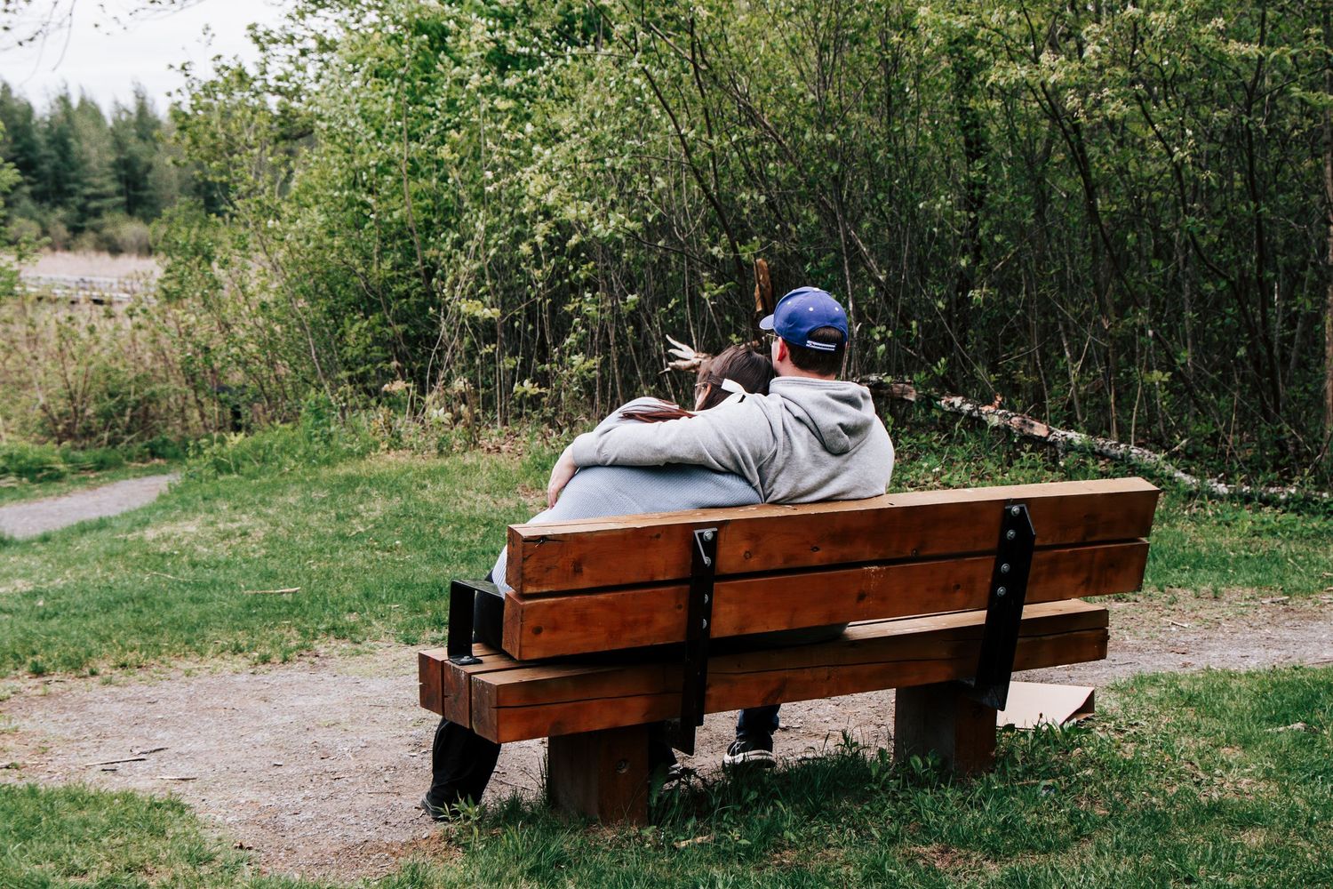 A person in a baseball cap sits alone on a wooden bench overlooking a green forest path.