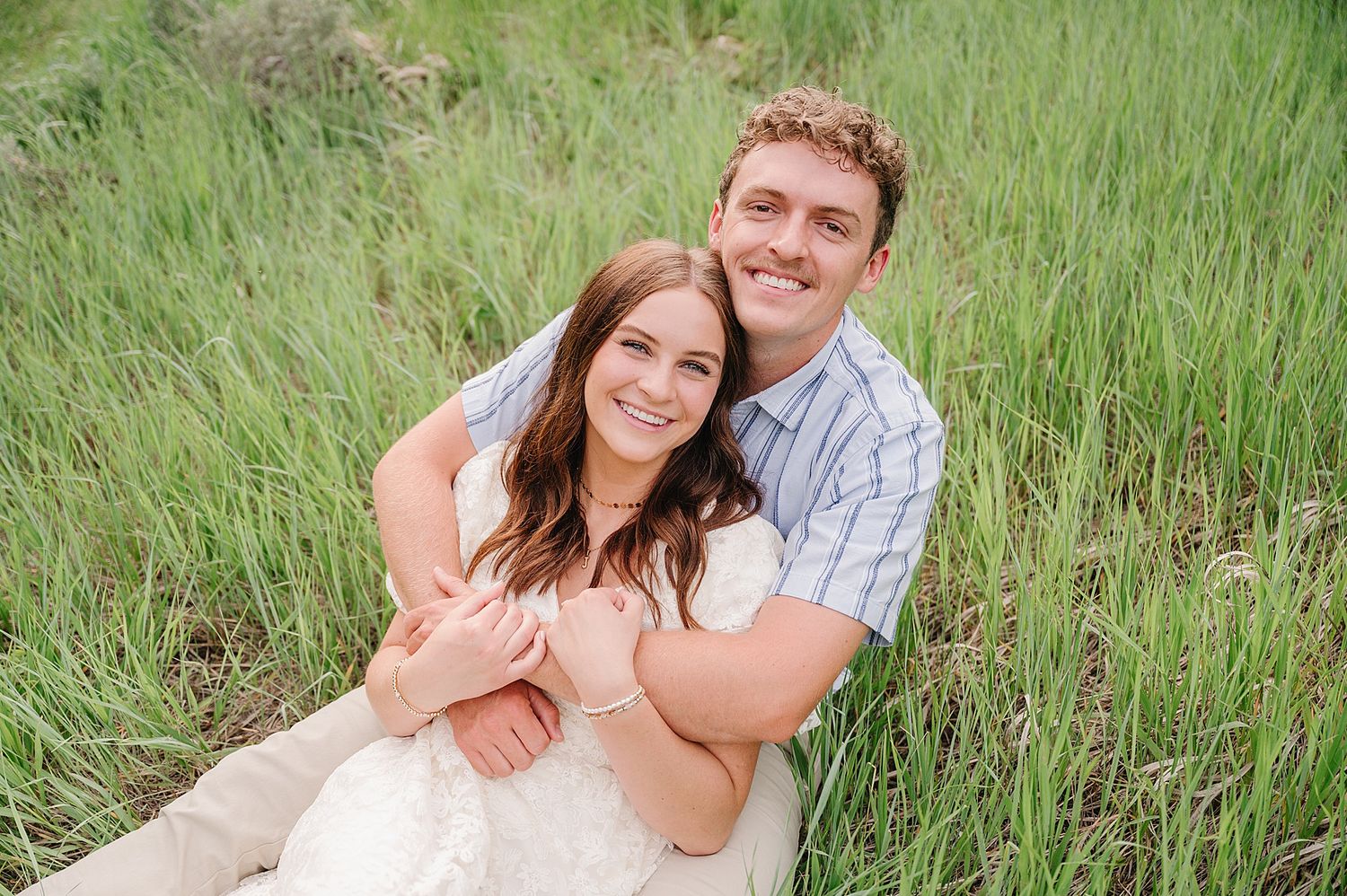 A happy couple embraces while sitting together in tall green grass on a sunny day.