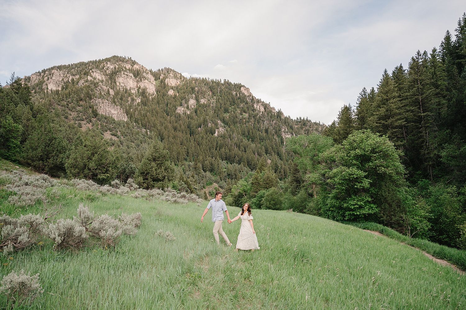 Two people walk hand in hand through a lush green meadow with majestic mountain peaks and evergreen forest in background.