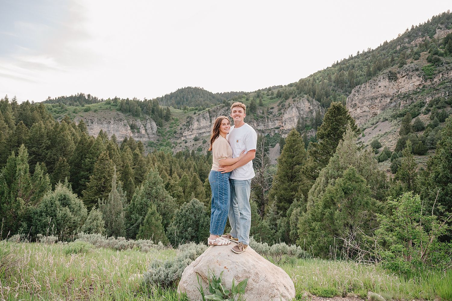 A couple stands on a large boulder surrounded by evergreen trees and rocky mountain cliffs in the background.