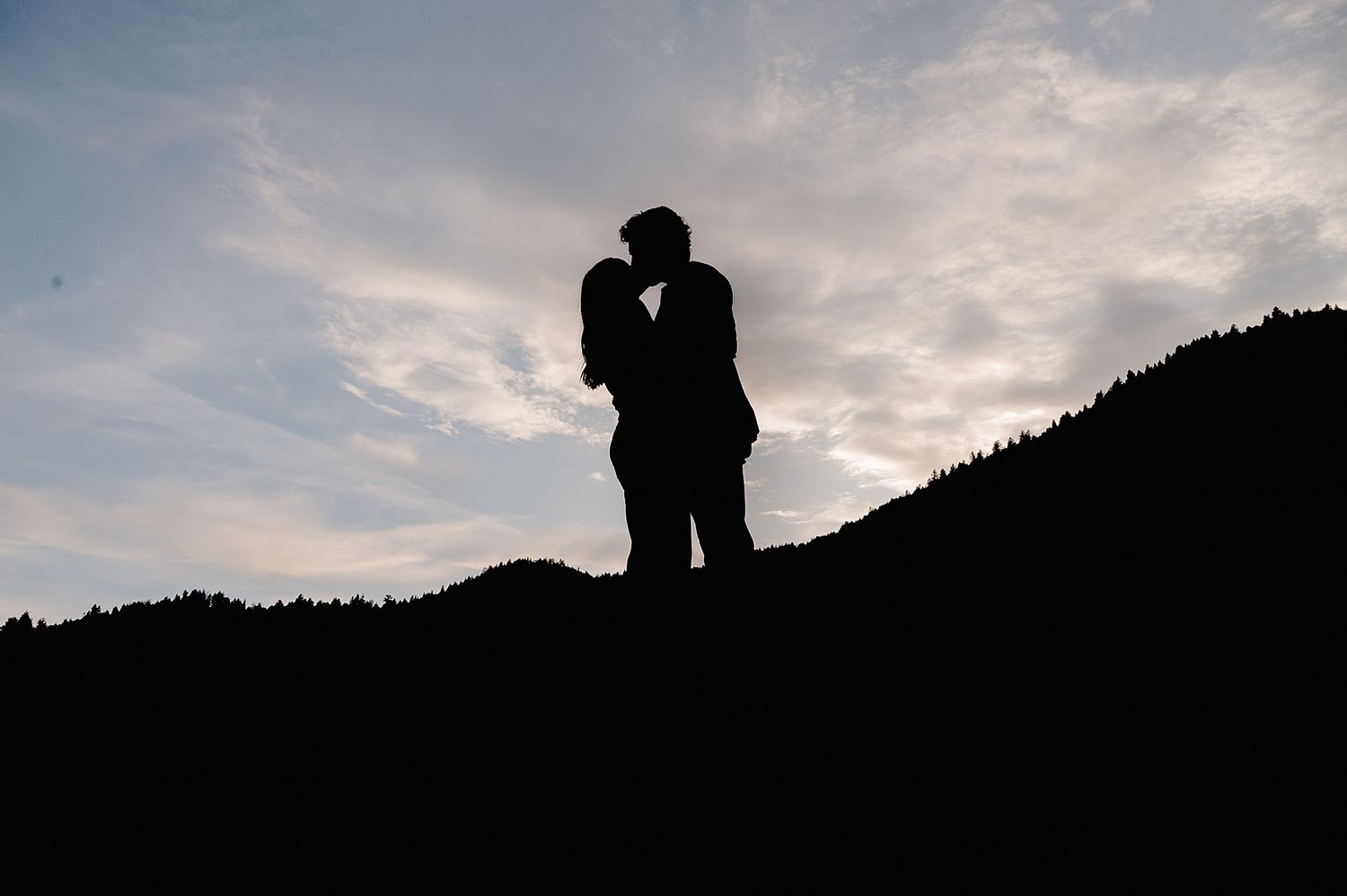 A silhouetted couple shares a romantic moment on a hilltop against a cloudy sunset sky.