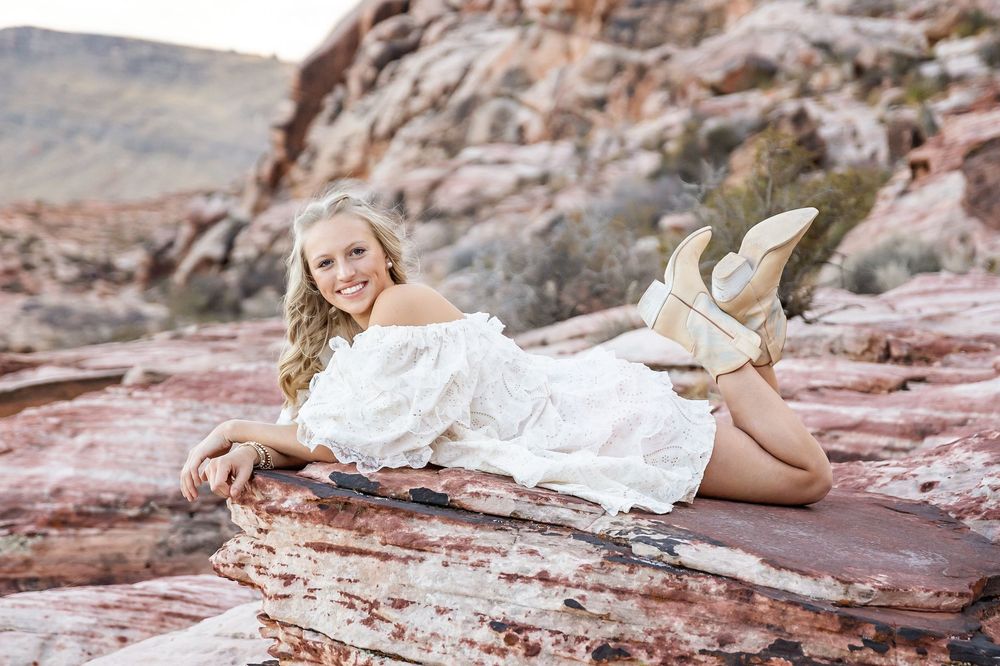 A person in a flowing white dress reclines gracefully on red rock formations in a desert landscape.