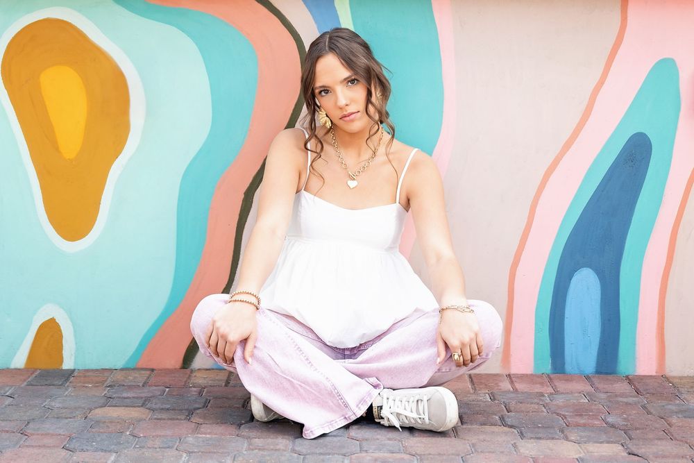 Someone sits cross-legged in a white tank top and pink pants against a colorful mural wall with wavy abstract patterns.