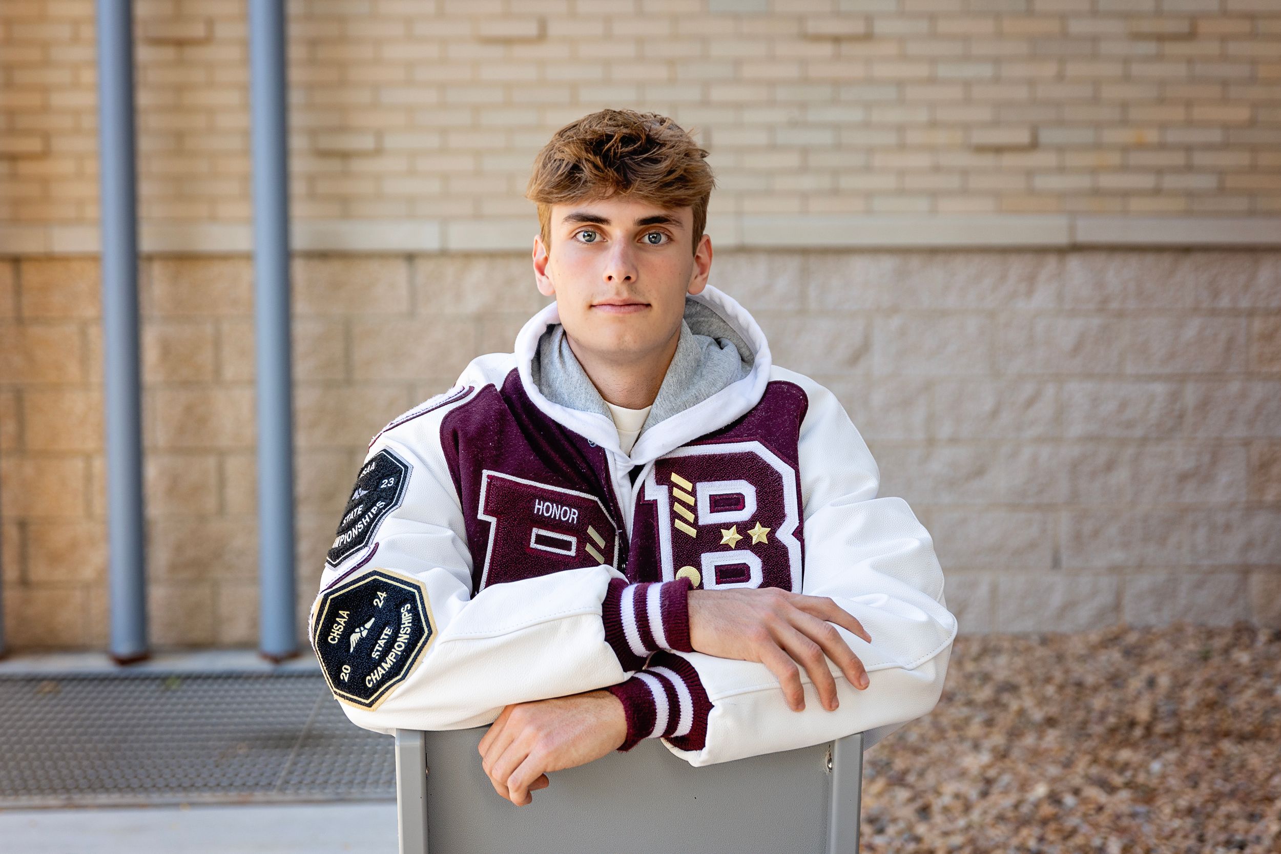 Student in maroon and white varsity letterman jacket leans against wall during outdoor portrait session.
