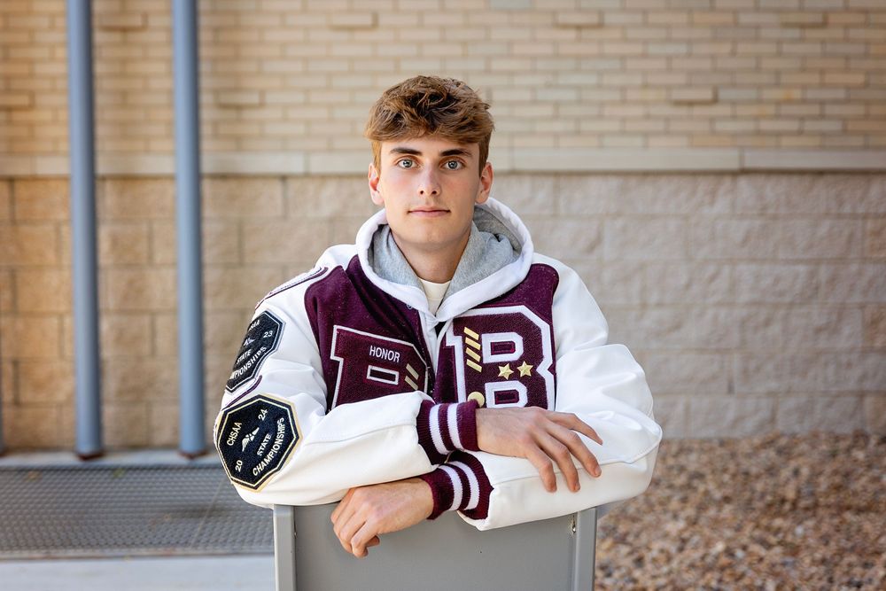 Student in maroon and white varsity letterman jacket leans against wall during outdoor portrait session.