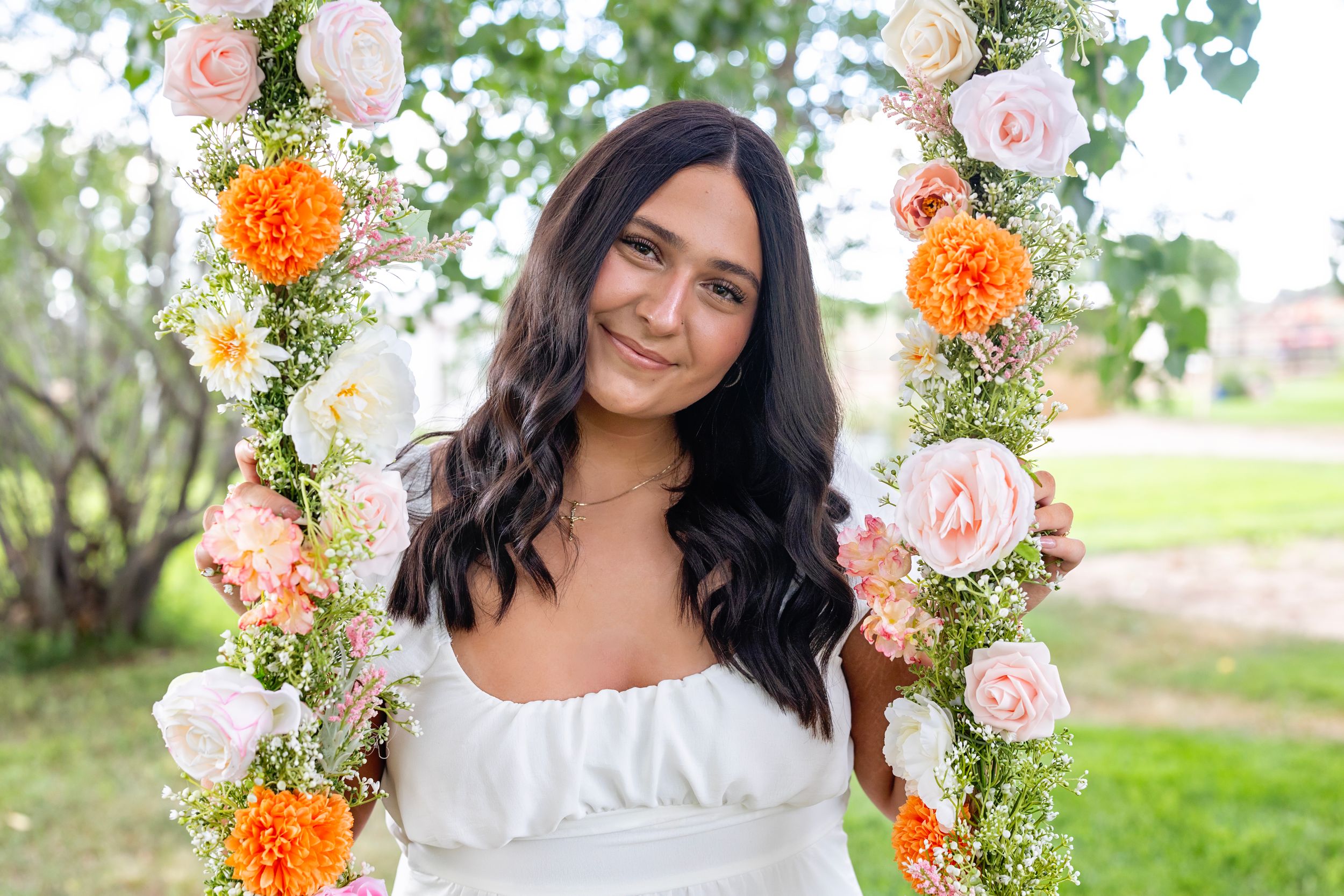 Someone poses on a flower swing decorated with pink and orange roses against a green outdoor background.