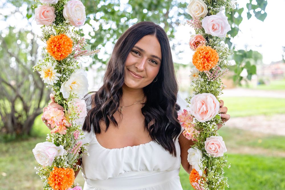 Someone poses on a flower swing decorated with pink and orange roses against a green outdoor background.