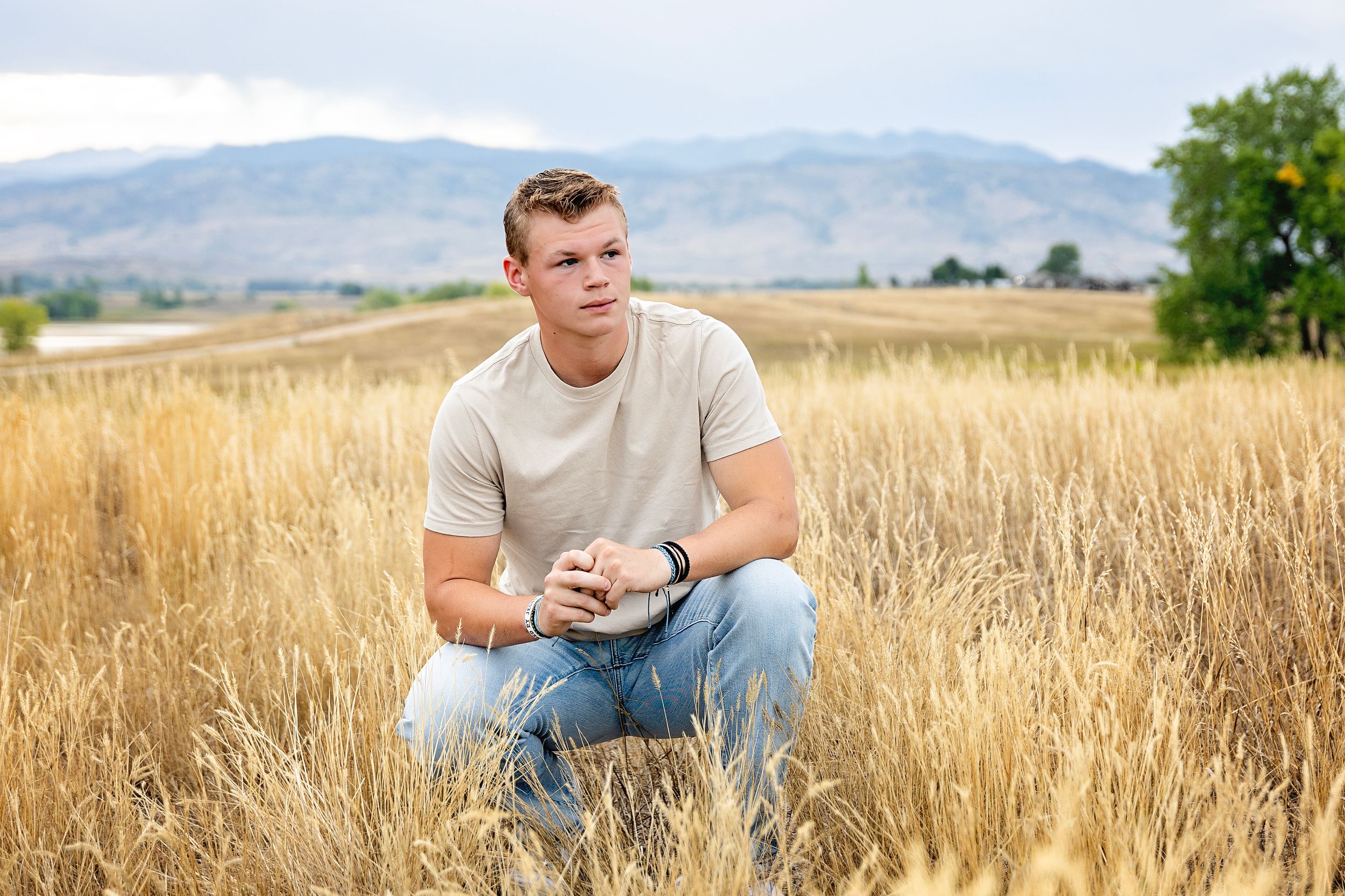 Person in white t-shirt and jeans crouches in golden wheat field with mountains in background.