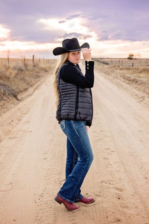 Person in puffy vest and cowboy hat poses on dirt road during sunset.