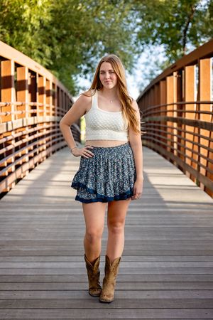 Person in crop top and floral skirt poses on wooden bridge wearing cowboy boots.