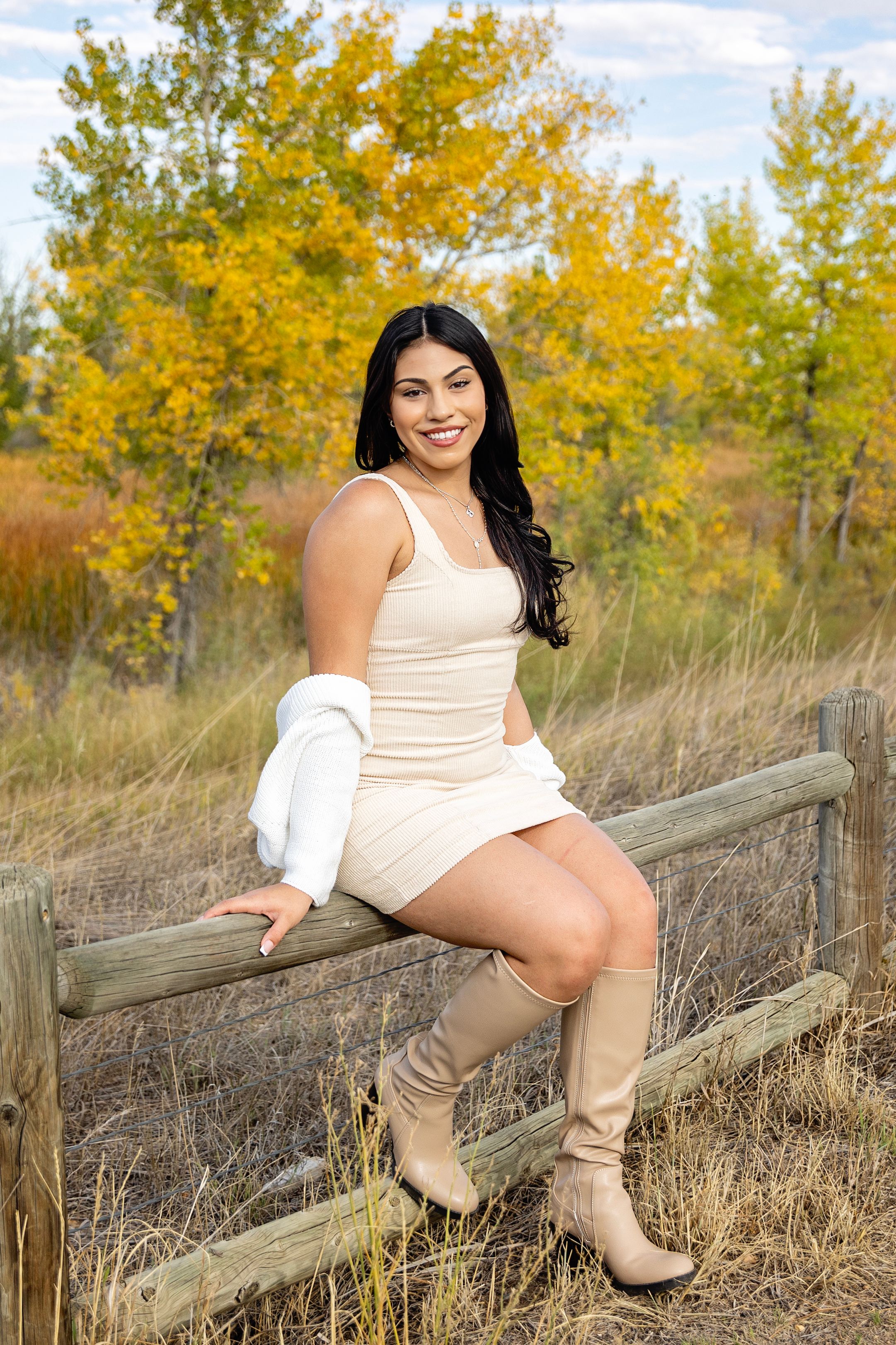 Person in cream dress and boots sits on wooden fence with autumn foliage in background.